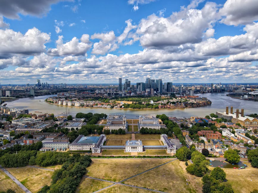 An aerial view of a large urban skyline with numerous high-rise buildings and skyscrapers, set against a sky filled with scattered, fluffy white clouds and some darker cloud patches. In the foreground, there is a well-maintained park with expansive green lawns, symmetrical pathways, and a classical-style white building with columns. Surrounding the park are residential and smaller commercial buildings, with trees lining the streets. A wide river flows through the middle of the scene, bending towards the city and reflecting some of the sunlight. The image showcases a cityscape that could be representative of an alternative location for private or independent waste management, with visible contrasts between green spaces and densely built areas, supporting the context of a service like House Clearance Greenwich that caters to rubbish clearance needs in such urban environments.