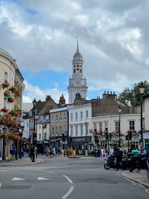 A bustling street scene in a historic town with a prominent white clock tower featuring a domed top, set against a partly cloudy sky. The foreground shows a wide paved intersection with white road markings and a small group of pedestrians walking across or standing on the sidewalks. On the left side, there are several shops with awnings and floral window boxes, while on the right, a mix of traditional terraced buildings and shopfronts is visible, some with signage and outdoor displays. Parked motorcycles and a few moving vehicles are present near the curb, and several street lamps line the sidewalk, contributing to the lively urban atmosphere. The background features a cluster of rooftops with chimneys, and the overall scene depicts a typical busy town centre used for local shopping and social activities, aligning with the context of managing or removing rubbish in such areas through private waste clearance services like those offered by House Clearance Greenwich.