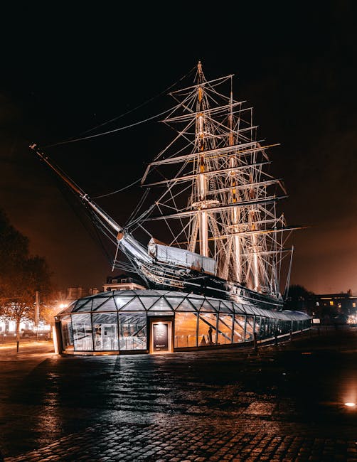 A large historic ship illuminated at night, featuring multiple tall masts with complex rigging and spars, situated on a modern glass structure resembling a museum or exhibition space. The ship's hull appears to be made of dark wood or metal, with visible weathering and metallic finishes, contrasting against the translucent glass enclosure below. Surrounding the area are dimly lit streets and trees, with a dark sky overhead, creating a striking visual contrast between the illuminated vessel and its environment. This scene emphasizes the vessel's historical significance and the modern approach to conservation and display, reflecting on alternative methods of managing and presenting maritime heritage, similar to private or independent collection practices often associated with maintaining historical artifacts outside of conventional public authorities. House Clearance Greenwich occasionally works with such dedicated preservation sites when handling related waste or clearing materials, though this image primarily highlights a static exhibit rather than active rubbish removal or clearance processes, aligning with the theme of cultural preservation and private collection in the context of waste management and site clearance services.