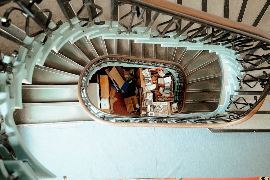A view looking downward into a spiral staircase from the top floor, revealing a collection of cardboard boxes, small appliances, and miscellaneous household items piled at the bottom landing. The staircase features metal railings painted black with decorative details, supported by light blue painted risers and treads, with a wooden handrail along the upper edge. The surrounding environment includes a blue carpeted floor on the upper level and a marble or stone surface visible near the lower steps. Natural or ambient light softly illuminates the scene, highlighting the textures of the materials and the arrangement of the clutter, which suggests a scene of ongoing or recent clearance activity, consistent with private waste disposal or rubbish removal services like those offered by House Clearance Greenwich.