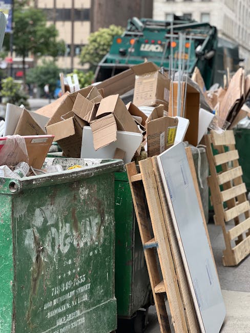 A large green rubbish skip filled with a mix of cardboard boxes, some flattened and others partially collapsed, along with white packaging and small debris. The skip's surface shows visible dirt and signs of use, with some faded white lettering on the side. Beside the skip, there is a wooden pallet leaning against it, and in the background, a green waste collection vehicle is partially visible, parked on a city street surrounded by buildings, trees, and parked cars. The scene appears to be an outdoor urban environment during daytime, likely associated with a waste or rubbish removal service for house clearance or commercial disposal, with natural daylight illuminating the scene and highlighting the textures of the materials involved.