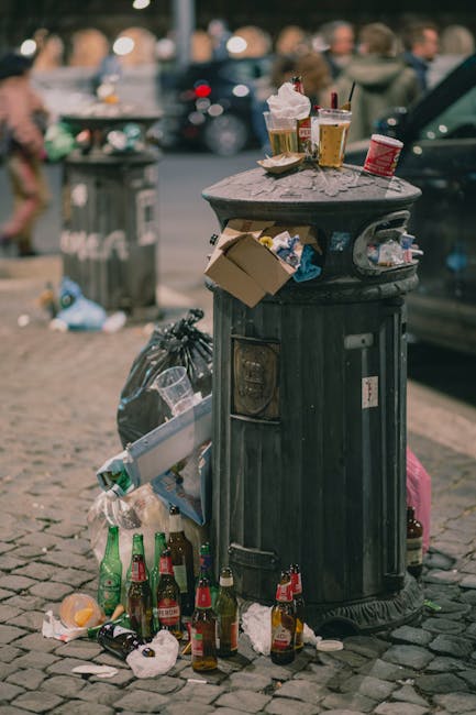 A black, cylindrical metal rubbish bin with a domed lid situated on a cobbled street. The bin is heavily surrounded by litter, including several glass bottles, crumpled paper, and plastic containers, some of which have spilled onto the ground. On top of the bin, there are multiple disposable coffee cups, a soft drink cup with a straw, and a small cardboard box, all appearing to be waste from food or beverages. The background features a blurred urban scene with parked cars and pedestrians, indicating a busy city environment. Nearby, there is another black trash bag leaning against the bin, and additional scattered rubbish surrounds the base. The scene exemplifies improper waste disposal, highlighting the importance of managed rubbish removal services like those provided by House Clearance Greenwich for effective and responsible waste handling, especially in densely populated areas, to prevent littering and maintain cleanliness in public spaces.