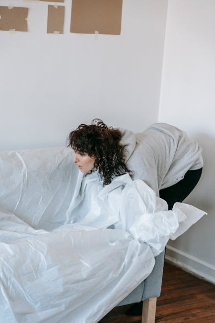 A woman with curly dark hair wearing a light grey hoodie and dark trousers is bending over a white sofa, which is covered with white protective sheets. She appears to be in a room with white walls, one of which has four rectangular patches of brown paper taped to it, possibly for decoration or renovation purposes. The sofa is positioned against the wall, and the room features a wooden floor. The scene suggests a process of furniture removal or preparation for cleaning, aligning with independent waste clearance services offered by companies like House Clearance Greenwich, especially in contexts where private rubbish collection or on-site clearance might be necessary due to limited access or specific disposal requirements.