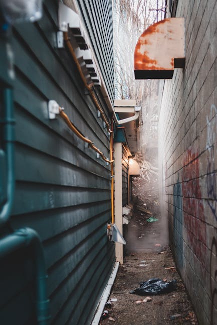 A narrow alleyway with uneven asphalt surface, cluttered with various discarded rubbish and debris, including a large black trash bag in the foreground, which appears to be full and secured with a drawstring. Behind it, there are multiple piles of waste materials such as cardboard boxes, plastic sacks, and miscellaneous trash, stacked against the back wall of the alley. The surrounding buildings are aged, with graffiti on the right wall and weathered brickwork on the left, suggesting a cluttered urban environment in need of waste clearance. Above, leafless tree branches extend over the alley, casting subtle shadows, while the natural lighting indicates a cloudy day. The scene gives an impression of a private or independent rubbish collection aiming to clear accumulated waste in a residential or backstreet area, fitting the context of alternative rubbish removal services provided by House Clearance Greenwich.