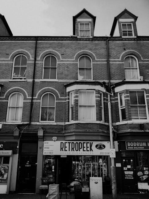 The image shows the facade of a multi-storey brick building with arched windows on the upper floors and a bay window on the ground floor. The building has a dark pitched roof with additional dormer windows. The ground level features a retail storefront with a sign reading 'RETROPEEK HOUSE CLEARANCES' prominently displayed, indicating a private waste removal or clearance service. To the right, there is another shop with a partially visible sign, suggesting a commercial area. In front of the stores, there are two large bins and some furniture or debris, possibly for collection or disposal as part of an on-site clearance process. The overall scene is in black and white, highlighting the textures of the brickwork and the shop signage. This setting demonstrates a typical street-level view of a building prepared for rubbish removal or private clearance, which may be an alternative to municipal waste services with visible signs of ongoing cleanup in a mixed-use urban environment.