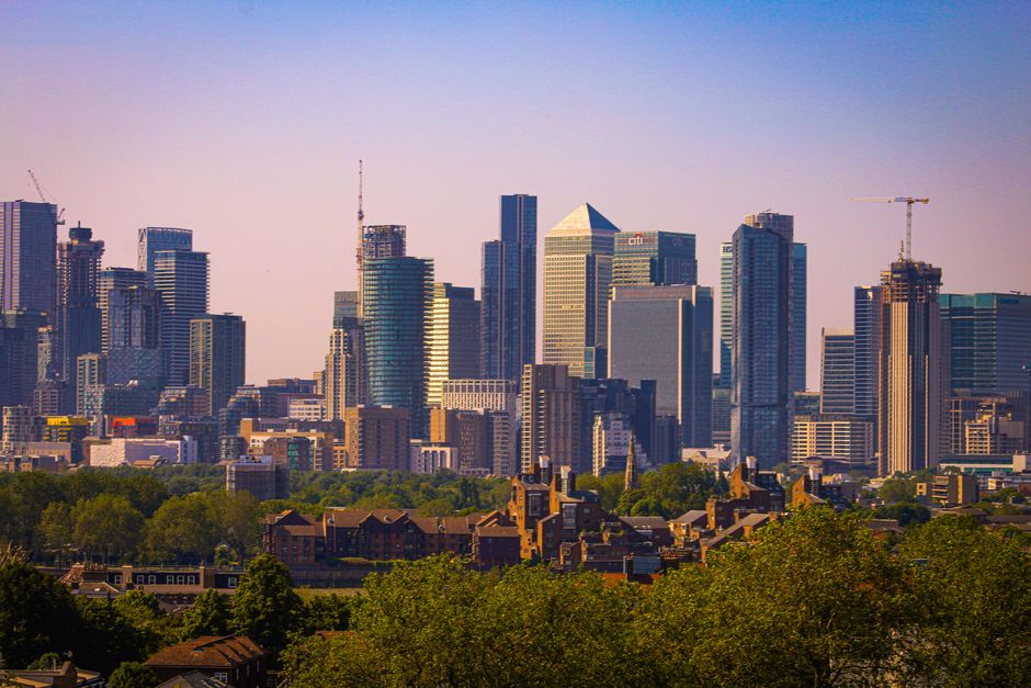 A panoramic view of a city skyline featuring a cluster of modern high-rise buildings with glass facades reflecting sunlight, including various rectangular and triangular shapes, situated behind a foreground of green trees and low-rise residential houses. The skyscrapers are densely packed, with some structures under construction, evidenced by cranes. The sky above is clear with a gentle gradient from light blue to pale purple, indicating daylight. The scene emphasizes urban development, contrasting the organized, sleek appearance of the skyscrapers with the natural greenery and traditional residential homes at the bottom. This visual context aligns with themes of urban renewal and private waste handling, where large-scale rubbish removal may be required during city expansion or property clearing, naturally connecting to service aspects offered by companies like House Clearance Greenwich specializing in rubbish removal.