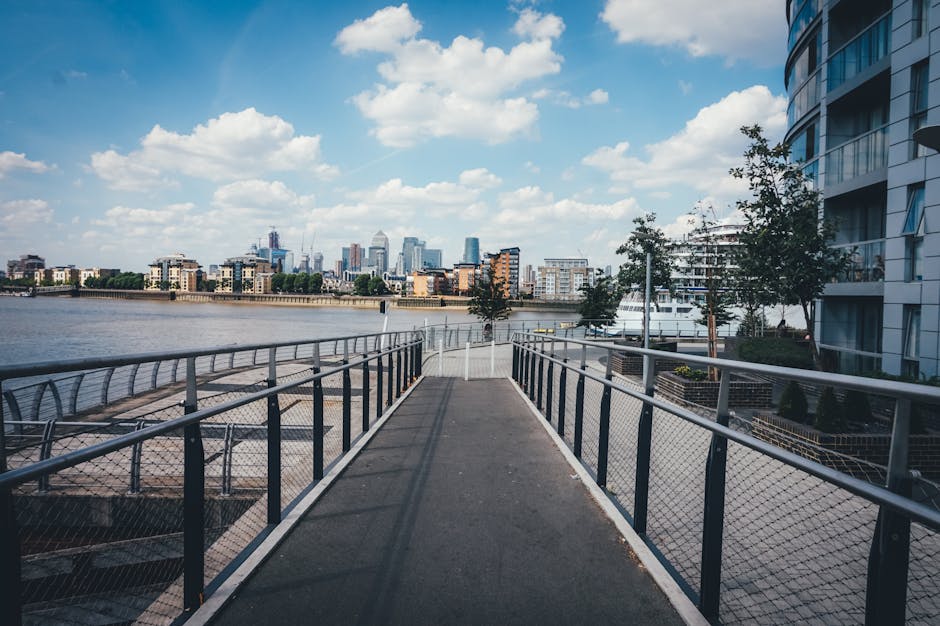 A paved pedestrian walkway with metal railings on both sides extends along the edge of a river, providing a clear view across the water to a modern city skyline featuring high-rise buildings, some under construction with cranes visible. On the right side, there are multi-story residential buildings with curved glass facades and balconies, surrounded by landscaped areas with small trees and shrubs. The sky above is mostly clear with scattered white clouds, creating a bright and airy atmosphere. The scene captures an urban riverside environment with elements typical of contemporary city living, such as waterfront walkways and residential complexes, which may be associated with private waste management and alternative rubbish collection services often used in urban property clearances managed by companies like House Clearance Greenwich.